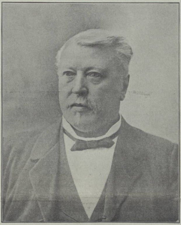 Portrait en noir et blanc d’un homme d’âge moyen. Il est montré en buste, portant un complet formel avec un gilet à motifs et un nœud papillon. Ses cheveux sont soigneusement coiffés et il a une moustache. Son expression est sérieuse et concentrée. 