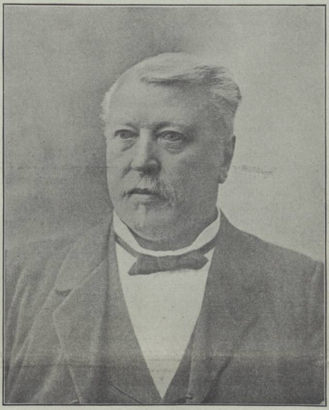 Portrait en noir et blanc d’un homme d’âge moyen. Il est montré en buste, portant un complet formel avec un gilet à motifs et un nœud papillon. Ses cheveux sont soigneusement coiffés et il a une moustache. Son expression est sérieuse et concentrée. 