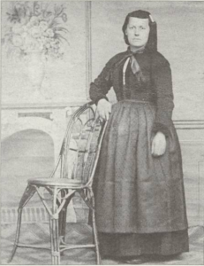 A full length black and white portrait of a woman standing with her arm resting on the back of a chair. She is wearing traditional Icelandic women's clothing, including a dark dress with an apron, a scarf tied around her neck, and a woolen cap with a tassel hanging down to her shoulder.