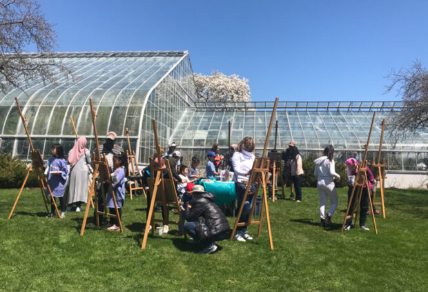 Photo of an outdoor painting workshop with children and adults painting at easels in a park. The scene is sunny with a greenhouse in the background.