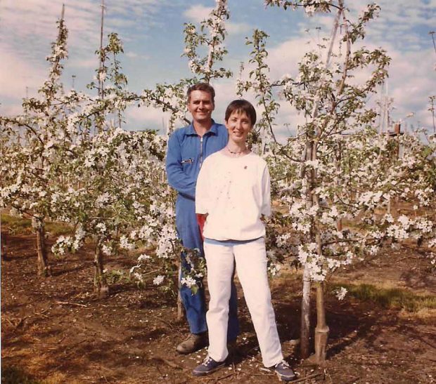 Colour photograph showing a woman and a man, Diane Goyette and Louis Poulin, standing in their orchard. The woman is dressed in white, the man in blue. Behind them, dwarf apple trees are visible.