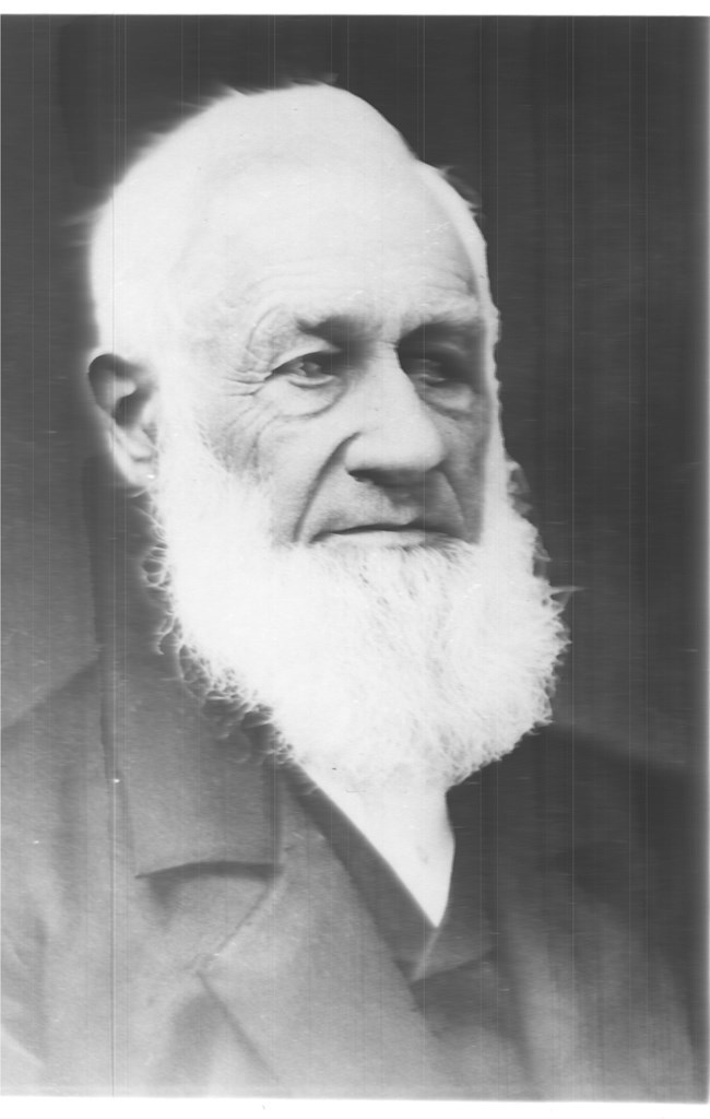 Black and white portrait photograph taken on an angle of Aaron Alexander Adams, with white hair and beard, dressed in a dark suit.