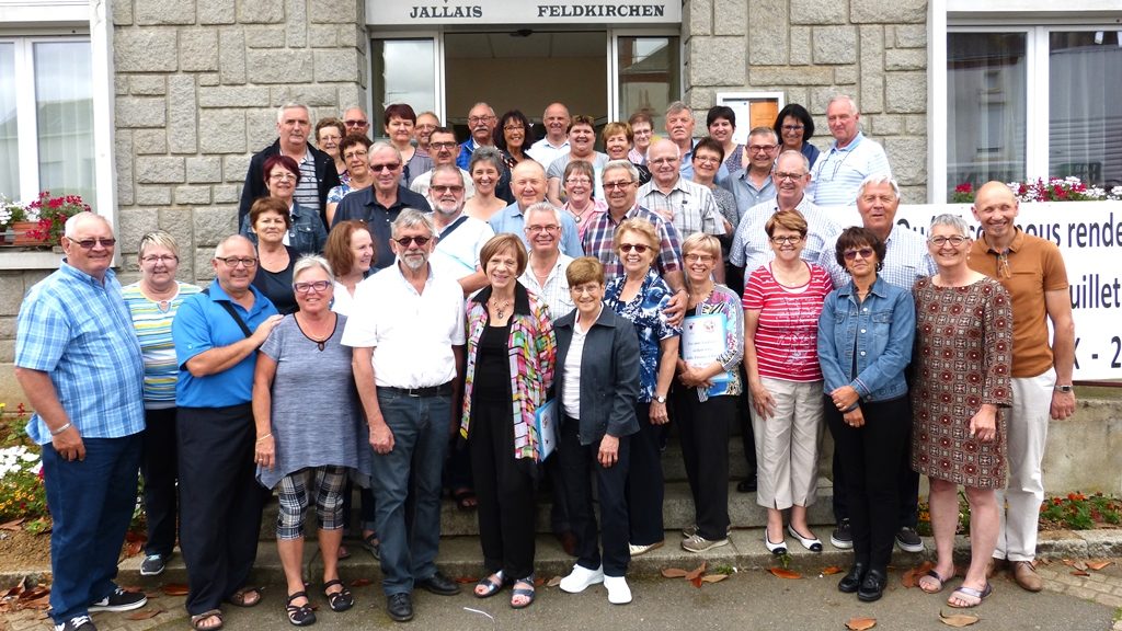 Colour image of a group of people standing in front of the municipal hall in Jallais, France. Dressed in summer clothing, they are gathered in front of the entranceway of a grey brick building.