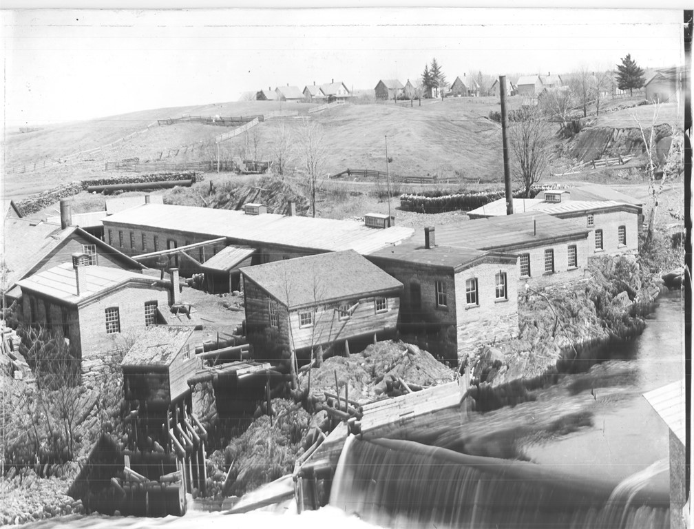 Black and white photograph of several factory buildings with falls from a dam on the Coaticook River in the foreground. In the distant background are some houses on the top of a hill.
