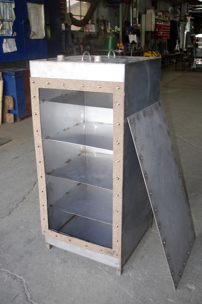 Colour photograph of a steel box in the workshop of the Lemieux steel company. The compartments of the capsule are visible, with its door leaning up against the capsule's right side, and a view of the workshop seen behind.