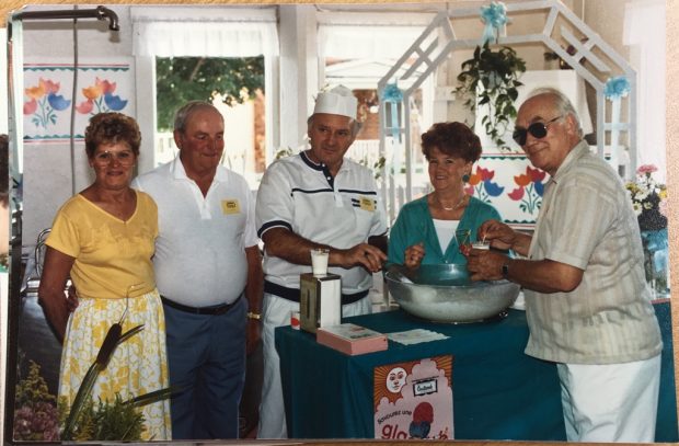 Colour photograph of, from left to right, Thérèse Houle, Fernand Houle, Émile Provencher, Gisèle Provencher, and Archbishop Jean-Marie Fortier. The group stands around a table covered by a teal-coloured tablecloth, on which stands a transparent bowl filled with the Vison blanc cocktail.