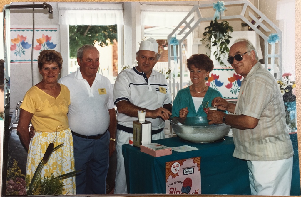 Colour photograph of, from left to right, Thérèse Houle, Fernand Houle, Émile Provencher, Gisèle Provencher, and Archbishop Jean-Marie Fortier. The group stands around a table covered by a teal-coloured tablecloth, on which stands a transparent bowl filled with the "Vison blanc" cocktail.