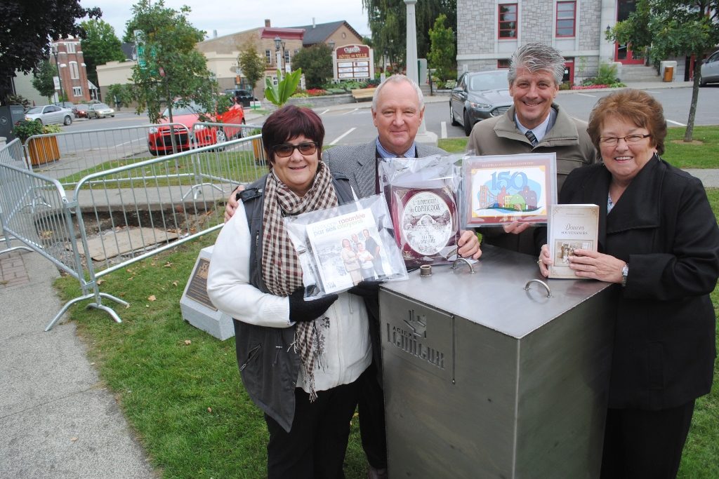 Colour photograph of four officials, two women and two men, posing beside the time capsule. They are holding documents produced for the 150th anniversary of Coaticook. Behind them is a partial view of downtown Coaticook.
