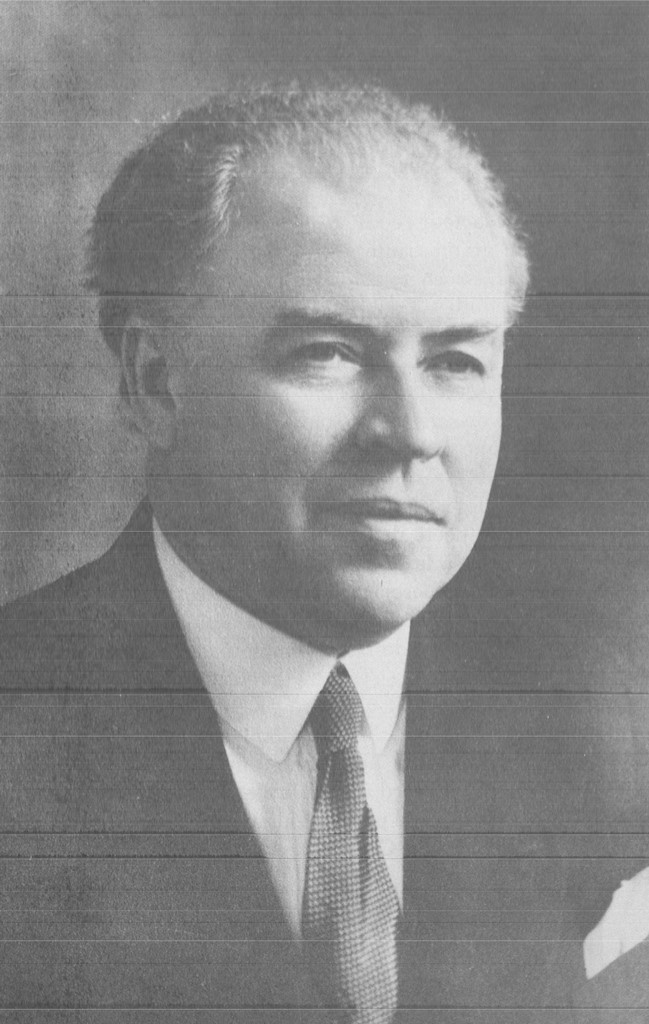 Black and white portrait photograph of Dr. Ernest Gendreau taken at an angle. He has grey hair and wears a dark suit, a white shirt and pocket square, and a white and grey tie.