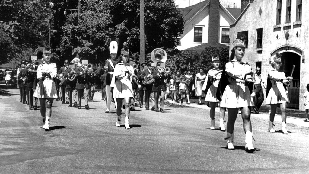 Black and white image of young women in white uniforms parading at the head of a band, made up of men in military uniform marching along Cutting Street in Coaticook, as spectators look on from the street, against a backdrop of trees and houses.
