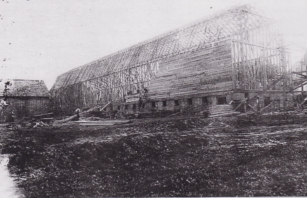 Black and white photograph of the frame of a very large structure under construction. Workers are beginning to attach boards to the frame.