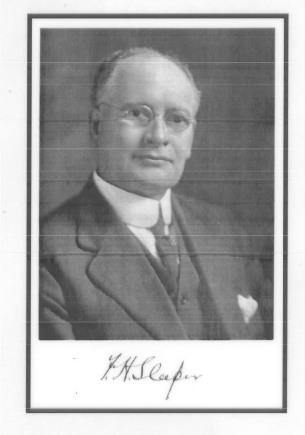 Black and white portrait photograph taken at a slight angle of Frank Sleeper wearing glasses, a dark suit and tie, and a white shirt and pocket square.