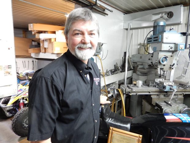 Colour portrait photograph of inventor Gilles Gagné, wearing a black work shirt and standing at a slight angle; he has grey hair, and a white beard and mustache. Behind him is a partial view of his workshop with machining tools and a storage section.