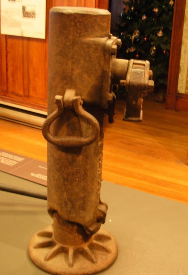 Colour photograph of a large, partially rusted tool on display in one of the Beaulne Museum's exhibition halls. Only a section of the floor and the wall of the hall is visible, as well as a Christmas tree located in a room further back.