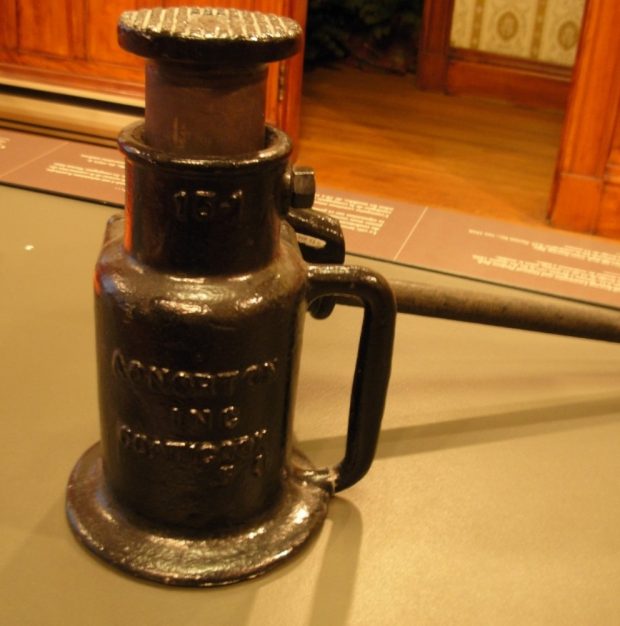 Colour photograph of a black tool on display in one of the Beaulne Museum's exhibition halls. Of relatively short length, it is inscribed with 15-1 at the top, and A.O. Norton Inc. Coaticook below. Behind it there is part of a wood-panelled wall, as well as a view of a a corridor leading further in the background.