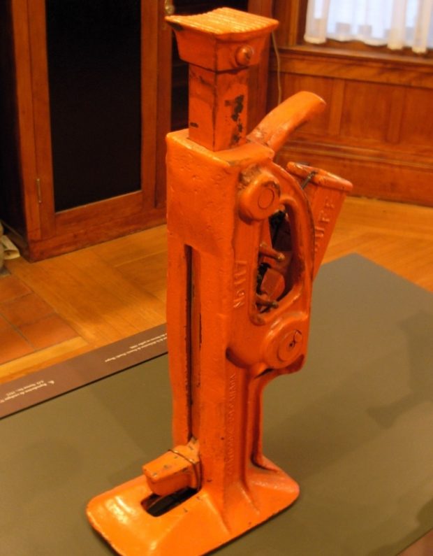Colour photograph of an orange-coloured tool on display in one of the Beaulne Museum's exhibition halls. Visible in the background is a cupboard and a section of the floor and wall.