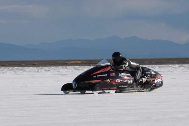 Colour photograph of a snowmobile being driven on a race course. The race is on the salt flats in the foreground; behind we see a stretch of desert land and a tall mountain range rises further back on the horizon.