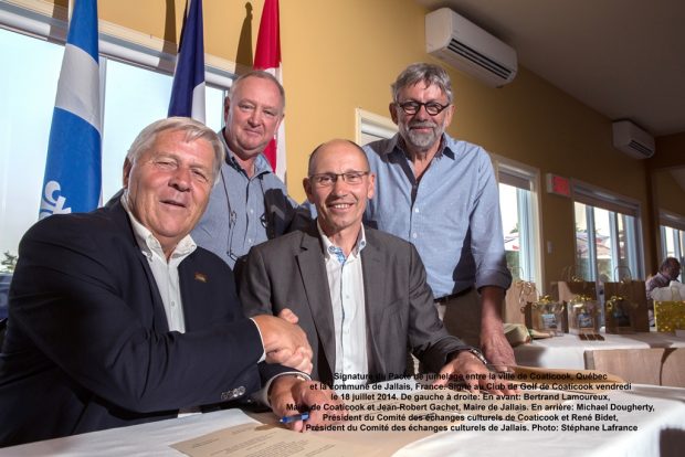 Colour photograph of a group of people: two men, wearing dark suits and white shirts, shake hands while seated; two others wearing blue shirts stand behind them. Behind the men, from left to right, are the flags of Québec, France and Canada. In the background to the right there are some wrapped gifts on tables.