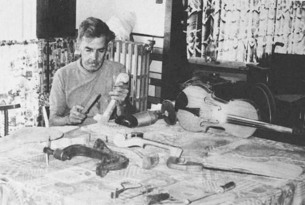 Black and white photograph of a man wearing a grey t-shirt, working at home, seated at a table. A violin, pieces of wood, and some tools lie on the table.