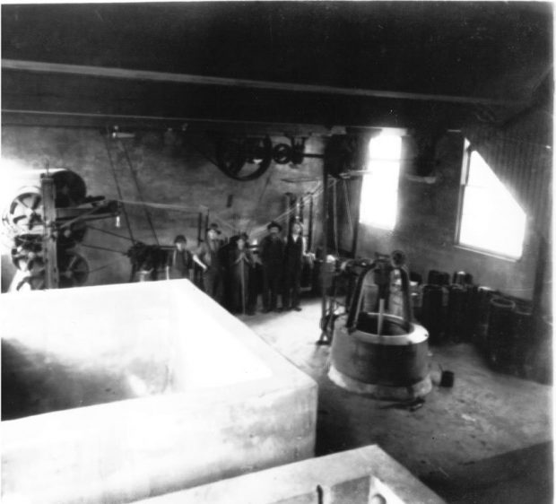 Black and white photograph of the interior of the factory, including machines, tools, materials and a few workers.