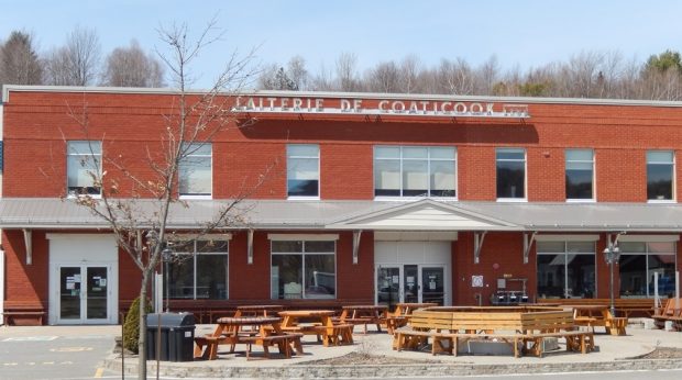 Colour photograph of the façade of a two-storey building, built mostly of red brick, with the tops of trees visible behind. There are two front entrances and many windows, wtih picnic tables and benches in front.