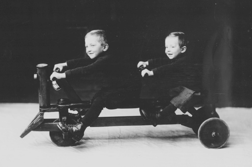 An old-fashioned black and white photograph of 2 smiling children seated on a cart that has 3 wheels and 2 sets of handlebars.