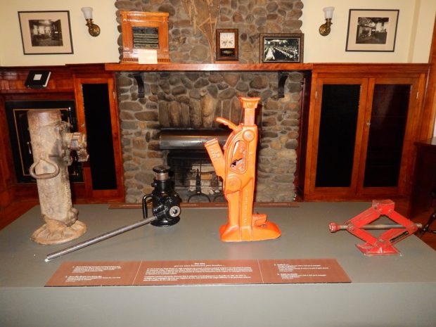 Colour photograph of four different Norton Jacks, displayed on a grey base in what used to be Mr. Norton's old office. Behind them is a wall with a stone fireplace in the middle, wooden cabinets on either side, and pictures hanging on the wall above.