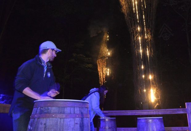 Colour photograph of two people beating drums, a man and a woman, with trees lit by special lighting in the background.