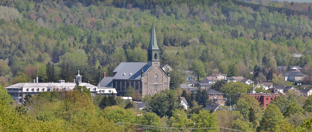Colour photograph of a view of Coaticook surrounded by forested areas. The church and steeple of St-Edmond are visible, as well as the large building of the school Collège Rivier along with a few other buildings.