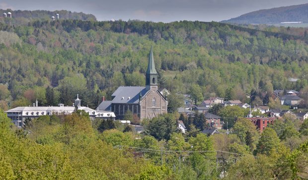 Colour photograph of a view of Coaticook surrounded by forested areas. The church and steeple of St-Edmond are visible, as well as the large building of the school Collège Rivier along with a few other buildings.