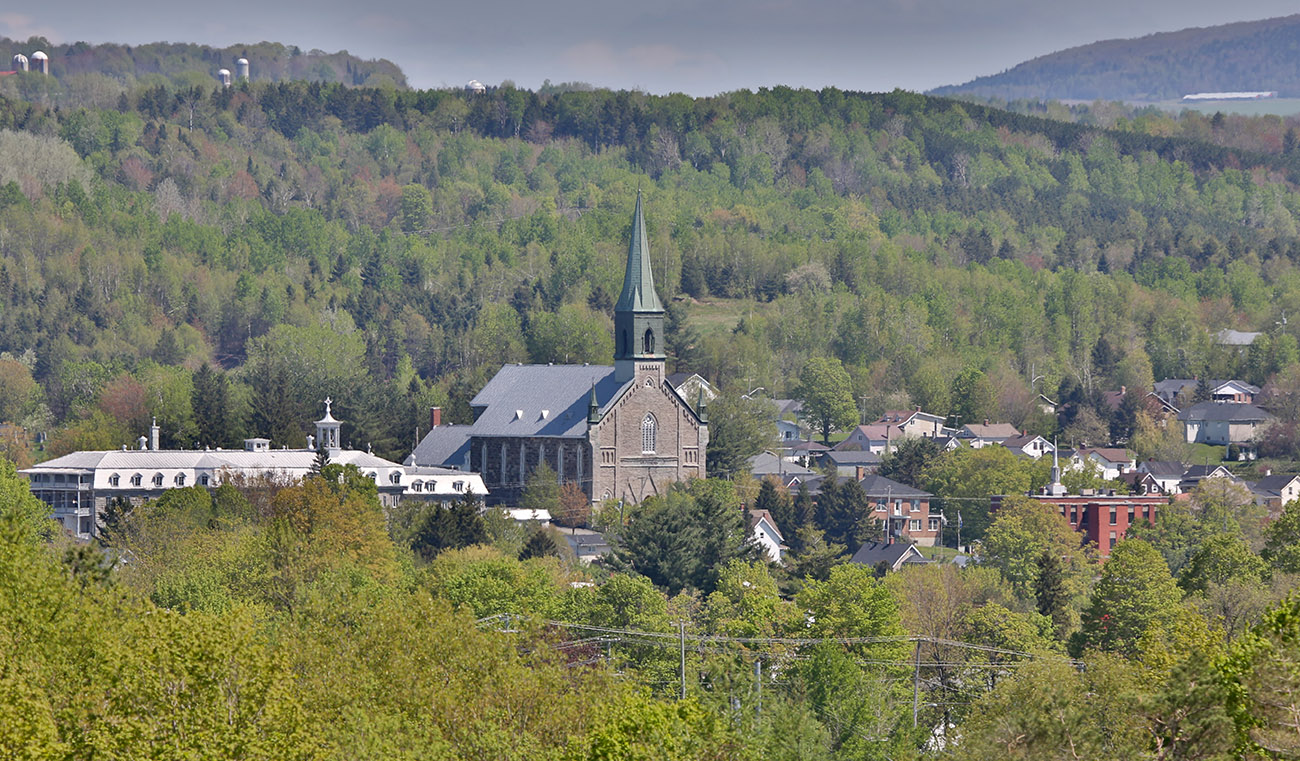 Colour photograph of a view of Coaticook surrounded by forested areas. The church and steeple of St-Edmond are visible, as well as the large building of the school "Collège Rivier" along with a few other buildings.