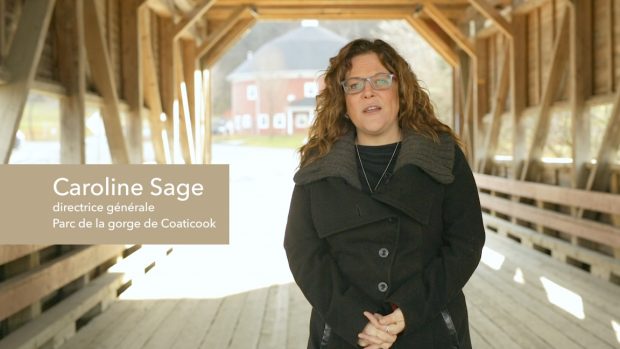 Colour photograph of Caroline Sage, the Director-General of the Coaticook Gorge Park, wearing a warm black coat. She's standing on the covered bridge of the park and behind her a round barn, another feature of the park, is visible.