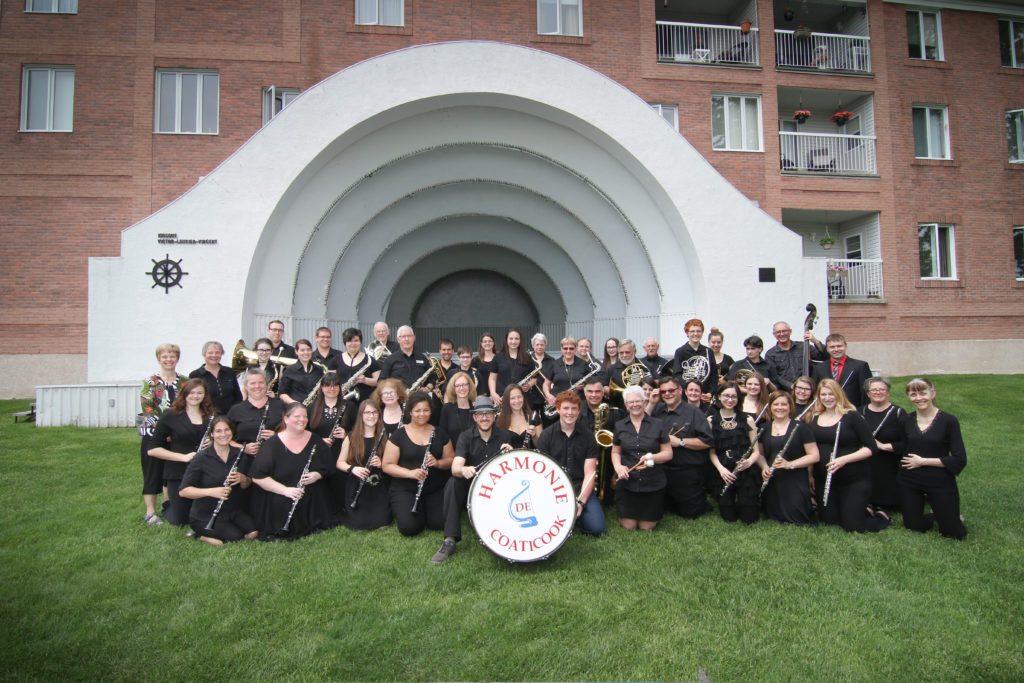 Photo couleur qui présente un groupe de musiciens de l'Harmonie de Coaticook tous vêtus de noir sauf une personne, la présidente. Ils se tiennent devant le kiosque de l'Harmonie de Coaticook qui est adossé à un bâtiment de brique rouge. 