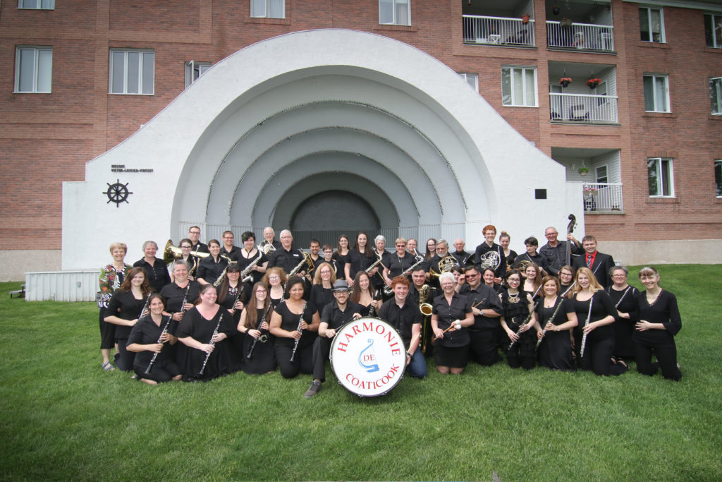 Colour photograph of a group of musicians from the Coaticook Harmony Band, all dressed in black except for the president, who wears a colourful outfit. They're standing in front of their outdoor concert stage, with a red brick apartment building in the background.