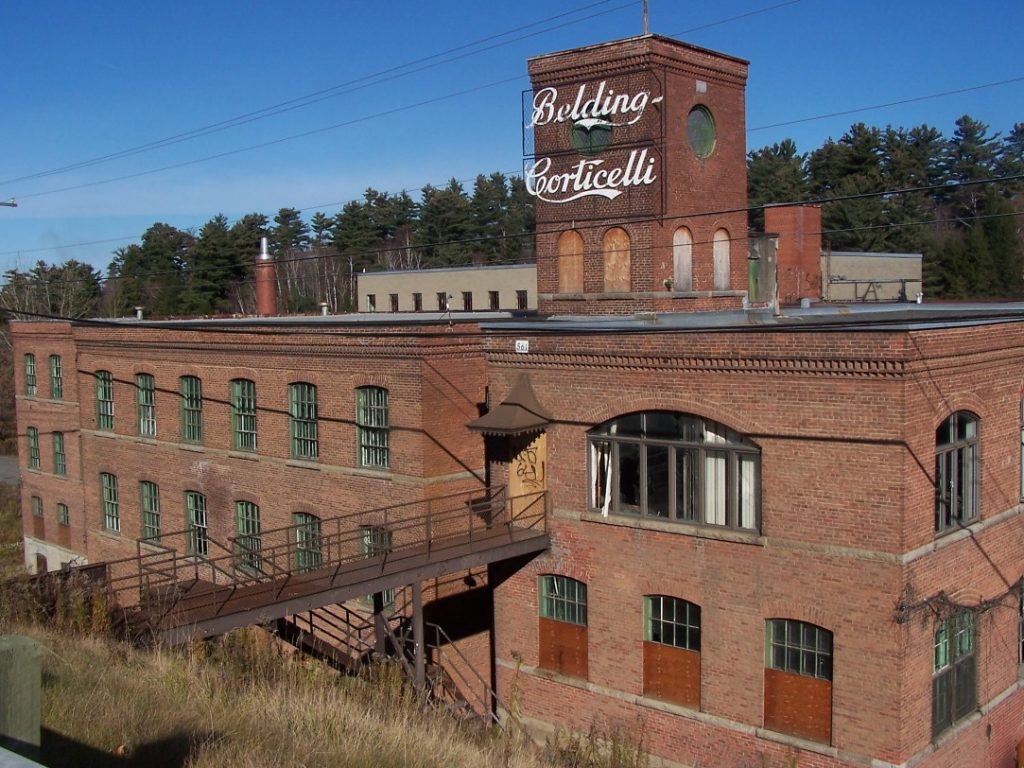 Colour photograph of the four-storey Belding Corticelli factory, built in red brick; a sign at the top of the building gives the name of the factory in stylized lettering. An iron walkway leads from the building to the road at left.