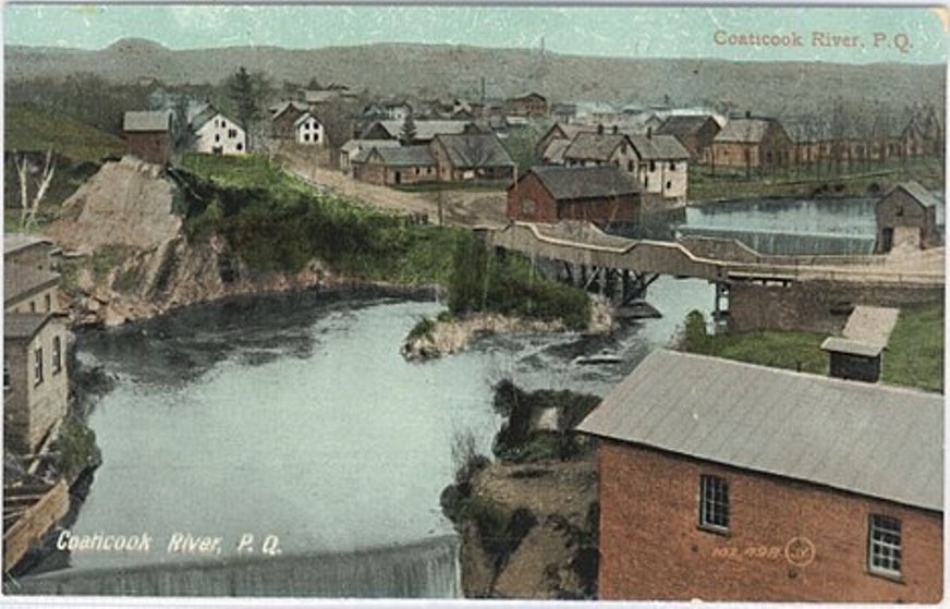 Historical colour photograph of an aerial view of the Coaticook River, showing a bridge and a few houses.