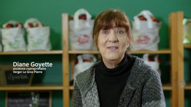 Colour photograph of Diane Goyette, co-owner of the orchard, Verger Le Gros Pierre, dressed in a black turtle neck and a grey cardigan. Behind her is a series of shelves containing apples in bags.