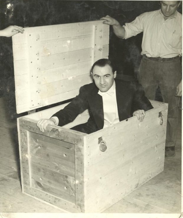 Black and white photograph of the magician “Grand Henri” seated in a wooden box, wearing a black suit, and a white shirt with a black bow tie. Standing to the right is a man dressed in dark pants and a light shirt, holding a lid for the box. Above to the left, can be seen someone's hand also gripping the box's lid.