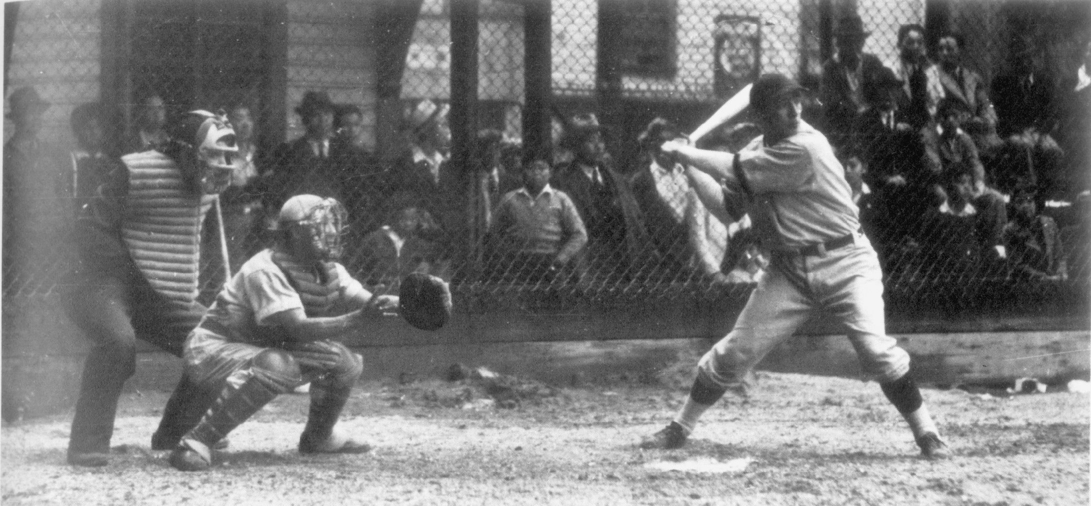 A man poised to hit a baseball with the umpire and catcher behind him with a group of people watching beyond a fence