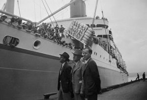 Three white men standing observing a ship filled with Japanese Canadians being exiled to Japan