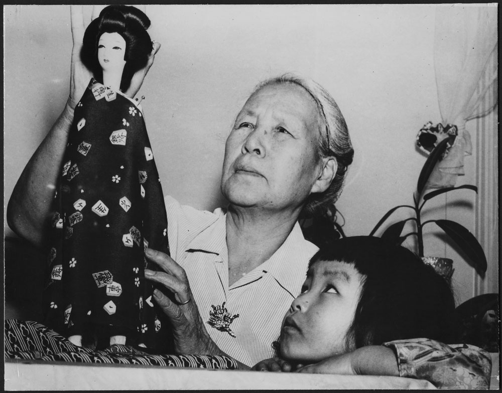 An elderly woman holding a doll dressed in Japanese fashion and a young girl observing