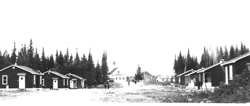 The first small houses located down the hill, near the lake. At the end of the road, we also see the church and the first presbytery. We see trees and people walking on the road where the houses are located on both sides.