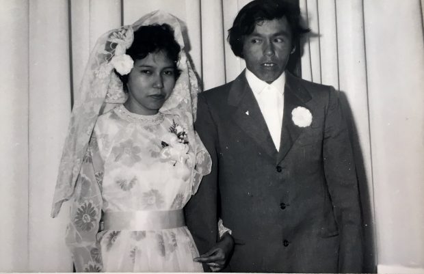 Anicinabek newlywed couple on their wedding day. The man wears a suit. The woman wears a white dress with flowers in addition to a veil in her hair and a flower girl. Black and white picture.