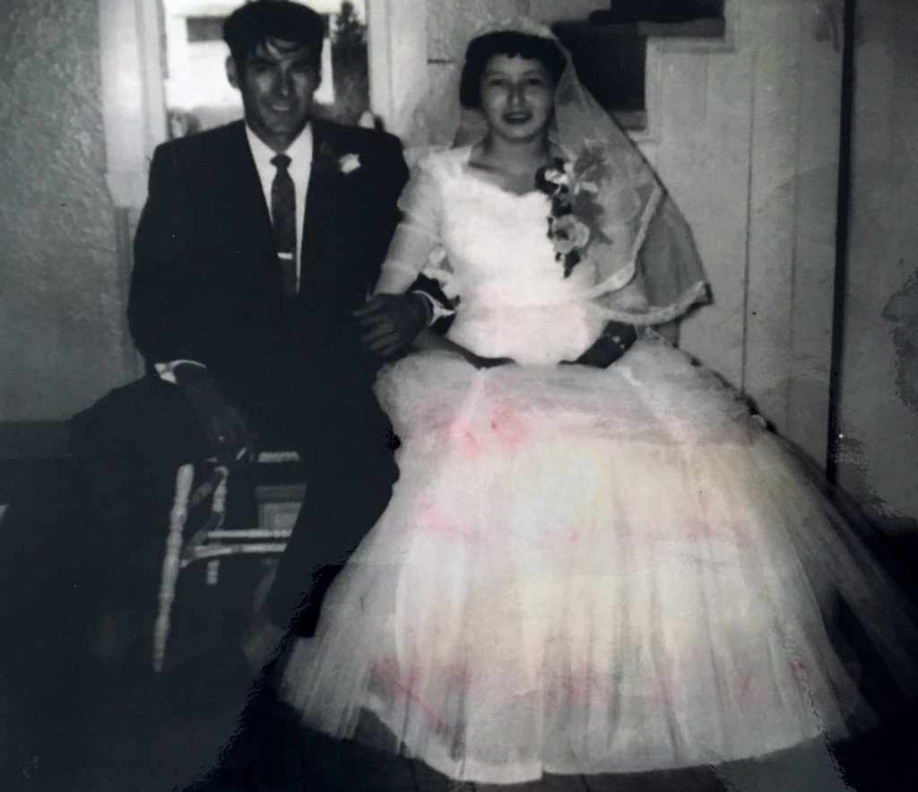 A young couple sitting side by side on their wedding day. The man is wearing a suit and tie and the woman is wearing a long tulle dress and a veil over her head. Black and white picture.