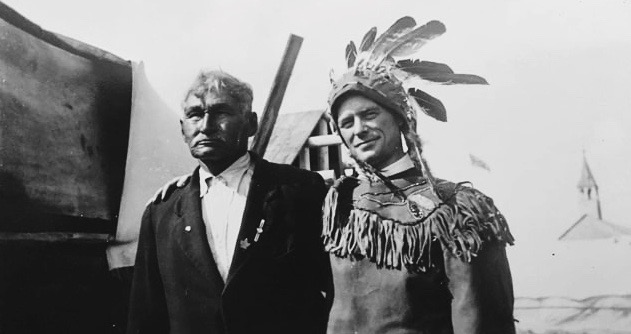 Grey Owl passing through Lac Simon with the leader of the band at the time. The chief is wearing a suit and Grey Owl is wearing a native headdress and a fringed skin suit. He is holding the chief by the shoulder. In the distance behind, we see a church steeple. Black and white picture.