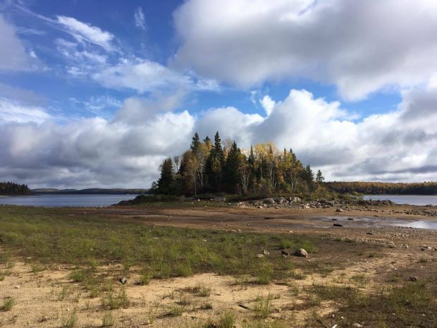 Fall landscape, Abitibi. A blue sky with white clouds. An island of coniferous and deciduous trees with yellow leaves. A lake in the vicinity with a dry portion (low tide) which lets see the ground of earth and rocks. Picture in color.