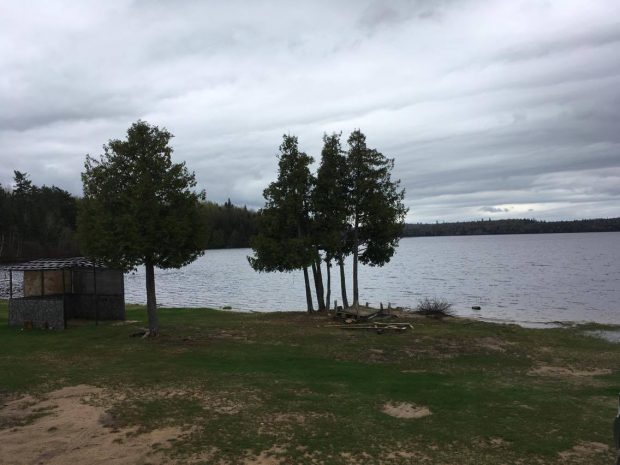 Cultural site of the community located at the waterfront. We can see some trees, a small portion of beach, the lake and the forest in the distance. The sky is dark. A small hut where they smoke meat is on the right. Picture in color.