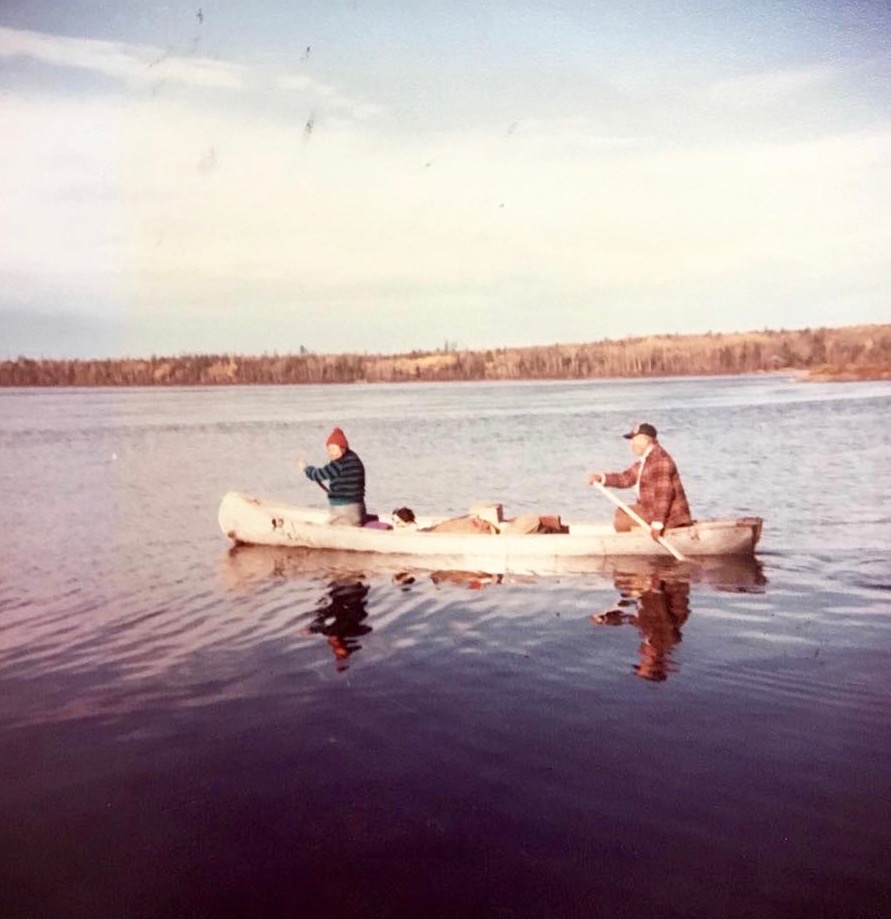 A couple travels by canoe on a lake. Both are paddling with the help of paddles. The man is in the back, the woman in the front. The forest is in the distance. Picture in color.