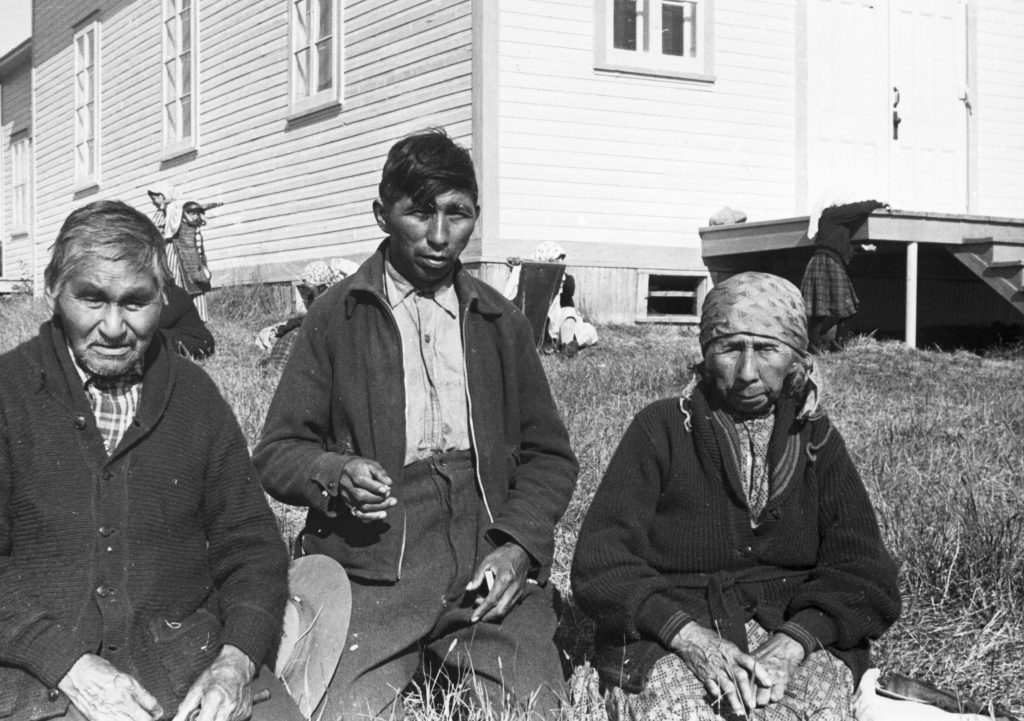 An Anicinabe family sits in the grass near Lac Simon. From left to right: an older man, a younger man (the son) and an older woman. They are wearing long clothes (jackets, shirts, pants, dress) and the woman has a scarf over her hair. The church is behind them. We also see children and a woman. Black and white picture.