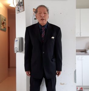Portrait of a former chief of Lac Simon. Dressed in a suit and tie, he stands in his kitchen. Picture in color.